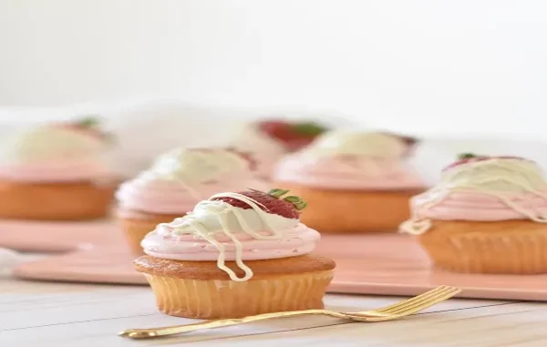 A close-up of a golden-brown cupcake with smooth pink frosting, topped with a white-chocolate-dipped strawberry and drizzle, with other cupcakes blurred in the background.