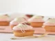 A close-up of a golden-brown cupcake with smooth pink frosting, topped with a white-chocolate-dipped strawberry and drizzle, with other cupcakes blurred in the background.