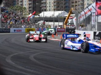 Multiple open-wheel race cars, including a blue and white number 3 car, speeding around a sharp turn on a street circuit track lined with spectators and sponsor banners.