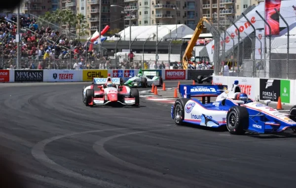 Multiple open-wheel race cars, including a blue and white number 3 car, speeding around a sharp turn on a street circuit track lined with spectators and sponsor banners.