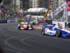 Multiple open-wheel race cars, including a blue and white number 3 car, speeding around a sharp turn on a street circuit track lined with spectators and sponsor banners.