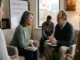 A diverse group of adults participating in a trauma-informed therapy session, sitting in a circle of armchairs in a bright, welcoming room. A middle-aged woman is speaking earnestly to an older male therapist holding a clipboard, while a whiteboard in the background lists "Understanding Trauma," "Coping Mechanisms," and "Recovery."