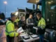 Two Department of Transportation (DOT) inspectors with a state trooper review logbooks, electronic tablets, and technical records at a roadside inspection table with a semi-truck at twilight.