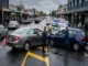 A police officer interviewing a frustrated driver at the scene of a two-car T-bone collision caused by a U-turn on a wet city street, with investigators reviewing the scene.