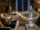 A realistic photo of a woman on a living room sofa looking at a realistic male hologram projected from a device on the coffee table. A glowing, golden infinity-shaped light stream flows between them, displaying floating text and icons of remembered details like "Your dog's name is Barnaby," "October 14th Anniversary," and "Recalling your interview last Tuesday."