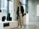 A person wearing a beige minimalist tracksuit under a structural, avant-garde black jacket in a contemporary art gallery.
