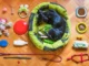 Flat lay arrangement of various pet products including a dog sleeping in a green bed, toys, food bowls, a leash, and grooming supplies on a wooden floor.