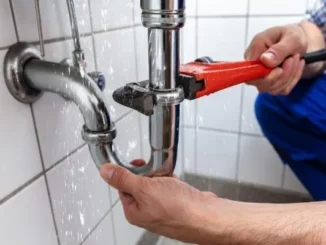 A plumber using a red pipe wrench to fix a spraying, leaking chrome p-trap under a sink.