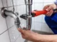 A plumber using a red pipe wrench to fix a spraying, leaking chrome p-trap under a sink.