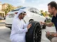 A man in a white Emirati thobe kneels on a stone driveway and inspects the deep tread of a new premium Michelin tire fitted to a white luxury SUV, while a smiling seller points to the brand name, in a sunny Dubai residential setting.