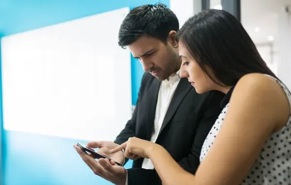 A man and a woman in an office setting looking closely at a smartphone screen, comparing and evaluating features to open a Demat account on a digital trading platform.