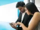 A man and a woman in an office setting looking closely at a smartphone screen, comparing and evaluating features to open a Demat account on a digital trading platform.