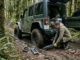 A person wearing work gloves kneels on a muddy forest trail to place a specialized metal vehicle recovery traction track in front of the tire of an off-road modified green Jeep Wrangler to gain traction and escape a rut.