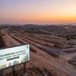 An aerial view of a large property development site during a sunset. A large billboard stands in the foreground, displaying information about DHA Gandhara Islamabad price prediction for 2026-2030, including a chart and illustrative elements of buildings. In the background, there are dirt roads and hills, with construction vehicles visible.