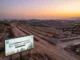 An aerial view of a large property development site during a sunset. A large billboard stands in the foreground, displaying information about DHA Gandhara Islamabad price prediction for 2026-2030, including a chart and illustrative elements of buildings. In the background, there are dirt roads and hills, with construction vehicles visible.