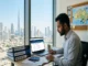 A realistic photograph of a male South Asian expatriate sitting at a desk in a high-rise Dubai apartment, filling out paperwork. His desk features a laptop displaying a points calculator, folders for Skills Assessment and IELTS prep, and a passport. The Burj Khalifa and Dubai skyline are visible through the window, and a map of Australia with "GOAL" sticky notes hangs on the wall.