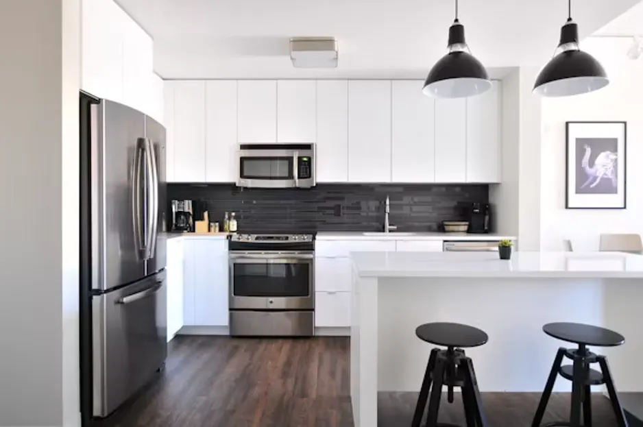 A contemporary kitchen featuring a full suite of stainless steel appliances, white cabinetry, dark backsplash, and a central island with stools.