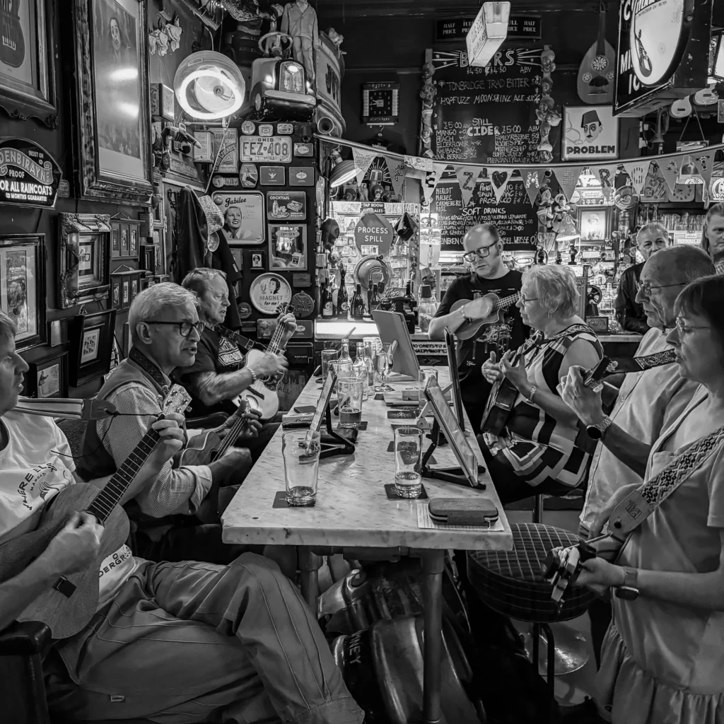 A black and white photograph of a group of older adults sitting around a long bar table playing ukuleles inside a traditional, eclectically decorated British pub.