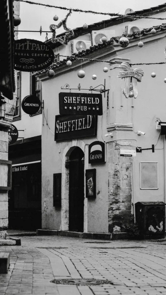 A black and white photograph of a narrow cobblestone street corner featuring the hanging wooden signs of "Sheffield Pub" under festive holiday garlands.