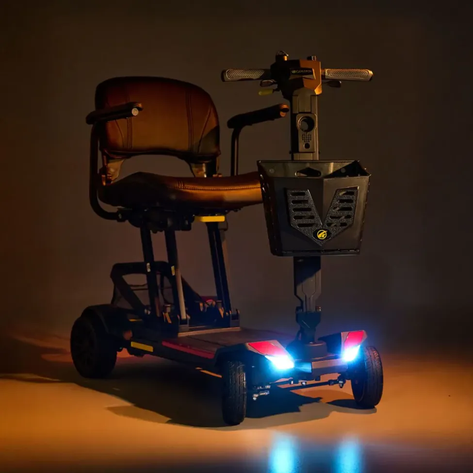 A compact black folding mobility scooter featuring padded seating, front basket, and illuminated LED headlights for travel and easy storage.