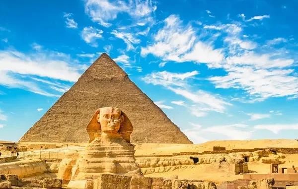 Front view of the Great Sphinx of Giza standing in front of the massive Pyramid of Khafre under a bright blue sky with wispy white clouds.