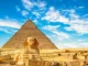 Front view of the Great Sphinx of Giza standing in front of the massive Pyramid of Khafre under a bright blue sky with wispy white clouds.