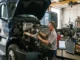 A focused mechanic, wearing safety glasses, performs maintenance on the engine and cooling system of a heavy-duty semi-truck in a busy repair bay, surrounded by parts and equipment.