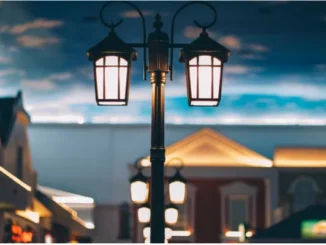 A double-lantern vintage style outdoor street lamp illuminated at twilight against a modern building backdrop.