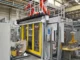 High-speed machine protection door with yellow borders and clear windows safeguarding an automated robotic work cell in an industrial manufacturing plant.
