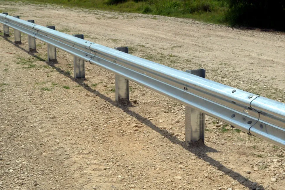 A newly installed galvanized steel W-beam highway guardrail running along a dirt roadside, designed for traffic safety and accident prevention.