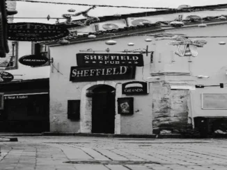 Black and white photograph of a historic European cobblestone street featuring the arched entryway and hanging signs of the "Sheffield Pub," adorned with festive overhead street decorations.