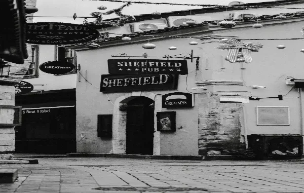 Black and white photograph of a historic European cobblestone street featuring the arched entryway and hanging signs of the "Sheffield Pub," adorned with festive overhead street decorations.