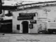 Black and white photograph of a historic European cobblestone street featuring the arched entryway and hanging signs of the "Sheffield Pub," adorned with festive overhead street decorations.