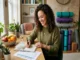 A smiling woman with curly hair sits at a wooden desk writing in a journal and reviewing a printed daily self-care routine tracker chart, surrounded by plants, a fruit bowl, and yoga mats in the background.