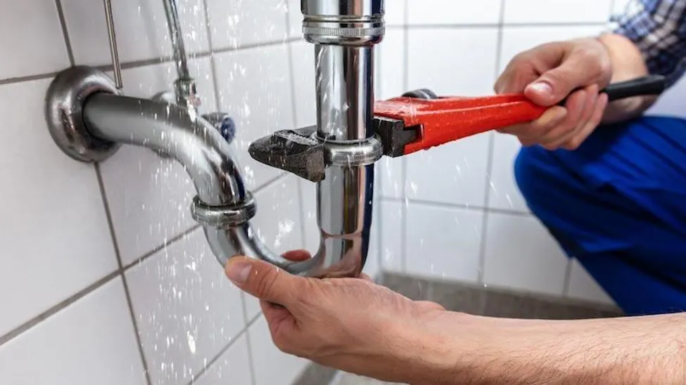 A close-up photograph of a plumber's hands using a large, red pipe wrench to tighten a Chrome P-trap connection under a sink, as water is spraying out from the joint in an old bathroom. The background shows white subway tiles, and the plumber is wearing a plaid shirt and blue work pants. This scene highlights the challenges of aging pipes in historic Brooklyn buildings.