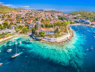 High aerial view of bright turquoise waters, boats, and red-roofed buildings surrounding the historic Franciscan Monastery in Hvar, Croatia.