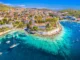 High aerial view of bright turquoise waters, boats, and red-roofed buildings surrounding the historic Franciscan Monastery in Hvar, Croatia.