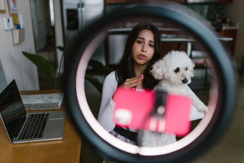 A female entrepreneur or content creator holding a small white dog while recording a video using a smartphone and ring light setup at home, representing digital marketing for a new pet product brand.
