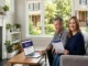 A smiling middle-aged couple sits on a living room sofa reviewing a Liberty Mutual home insurance policy document while a laptop on the coffee table displays a five-star review page.