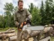 A man wearing safety glasses and gloves uses a chainsaw with an 18-inch guide bar to cut a log on a wooden sawhorse outdoors.