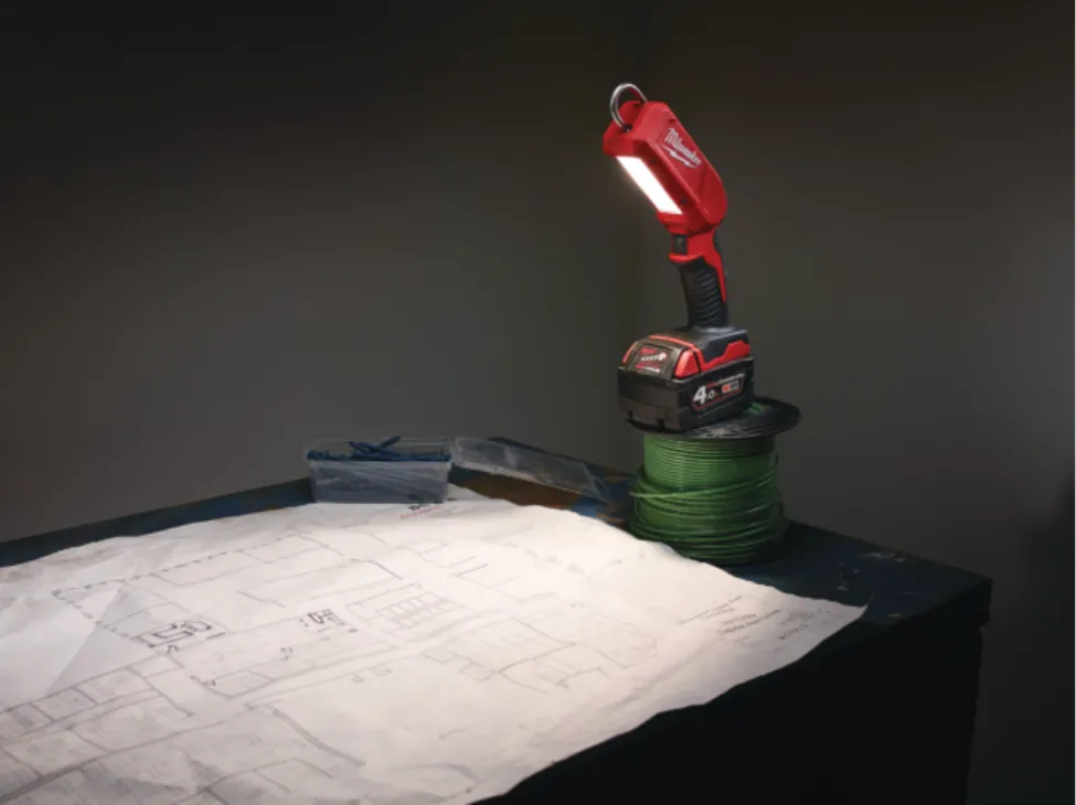 A cordless red Milwaukee LED work light illuminating architectural blueprints on a workbench, showcasing a leading competitor brand for a job site inspection light comparison.