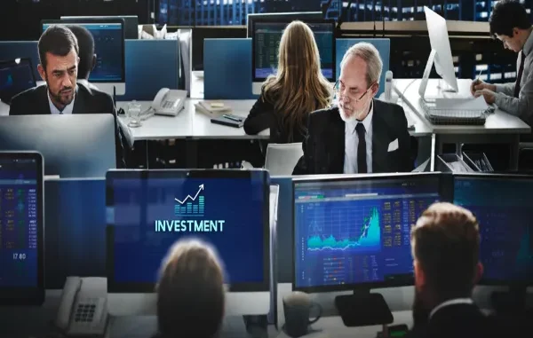 Financial traders working at desktop monitors displaying stock charts, data graphs, and an investment icon in a modern trading floor office.