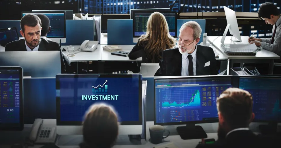 Diverse team of financial analysts and traders working at computer stations with stock charts and an "INVESTMENT" screen graphic in a modern trading floor setting.