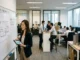 A woman in professional attire points to an office floor plan on a whiteboard, surrounded by other employees working in a modern office environment.
