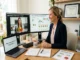 A smiling female nutrition consultant in a blazer sits at a bright wooden desk during a virtual meeting. Her dual-monitor setup shows a video call with a client holding fresh vegetables on one screen and custom meal-planning charts on the other. On the desk are a laptop, an open notebook with a meal plan sketch, and a printed plan titled "Flourishing Online Nutrition Practice."