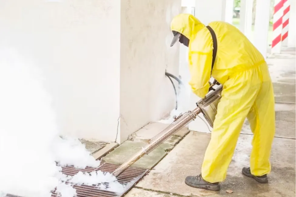 Pest control technician in a yellow protective suit spraying fog treatment along a home exterior drain.