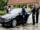 A candid photograph capturing a wedding couple boarding a black Mercedes-Benz S-Class sedan. A formally dressed chauffeur holds the door, with the groom helping the bride. The setting is a paved courtyard of a European venue in the Netherlands, with blurred VIP guests in the background. Natural daylight.