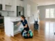 Woman with Queen City Cleaning Services scrubs the interior of a white cabinet shelf in a large, empty, modern open-concept kitchen with white cabinets, white subway tile backsplash, and hardwood floors and an empty living room in the background.