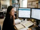 A focused female researcher sits at a multi-monitor workstation in a laboratory office, analyzing "Journal Analytics" metrics including SJR on a screen, with manuscripts and a "Scopus Indexed Journals" notice on the wall behind her.
