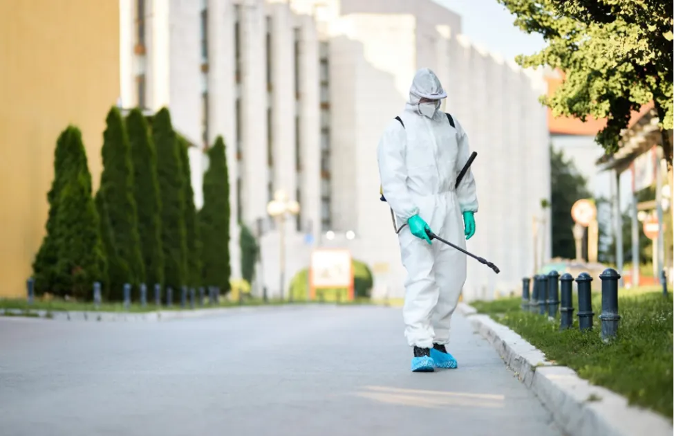 Professional pest control technician wearing full protective suit spraying pesticide outdoors to protect a building against seasonal pests.